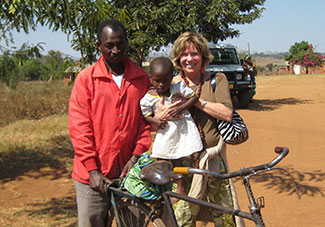 Peggy Bentley holds a young child, stands next to a man who balances a bicycle on a dusty road in Malawi