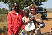 Peggy Bentley holds a young child, stands next to a man who balances a bicycle on a dusty road in Malawi