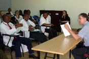 Students sit at desks in classroom, man sits behind table at front of classroom, addresses students