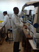 Man in white lab coat in laboratory lifts hood on complex equipment, woman in background works with similar equipment