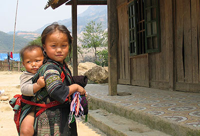 Young Vietnamese girl with boy toddler strapped to her back, standing in front of wooden house on dry dirt ground