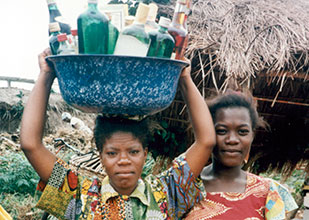 © 1996 Sara A. Holtz, Courtesy of Photoshare, Two African women outdoors, one balances tub full of alcohol bottles on her head