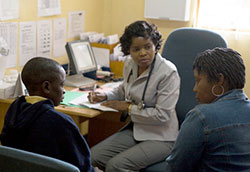 A medical professional takes notes during a discussion with a young child and a caregiver in a clinic.