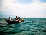 three young men in small wooden boat, fishing nets draped on side, wide expanse of blue water, mountains in distant background