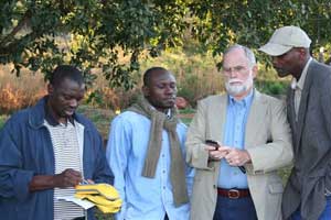 Man holds handheld mobile device, three other men look on, outdoors, trees in background