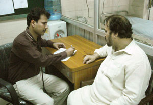 Man seated wearing stethoscope takes notes on piece of paper, man seated across from him, hospital bed in background