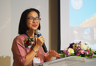 Dr. Waranuch Pitiphat, wearing a pink dress, speaks into a microphone while lecturing at a scientific conference. 