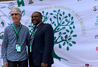In the photo on this page, Drs. Gregory Aarons and Eche Ezeanolue pose for a photo together in front of the white Nigeria Implementation Science Alliance banner. Aarons wears sunglasses, black pants and a blue striped shirt. Ezeanolue wears an open collar shirt beneath a dark suit.