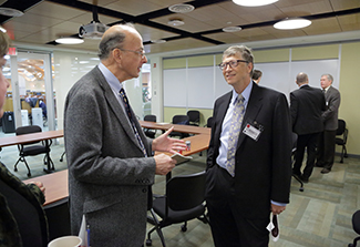 In the photograph, Dr. Roger Glass speaks informally with Bill Gates, founder of Microsoft and the Gates Foundation. They both wear suits and nametags and stand facing one another in a conference room.