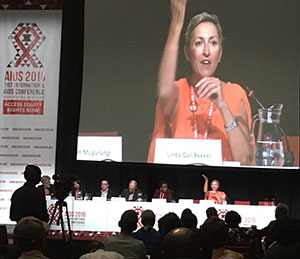 Linda-Gail Bekker speaks from a long table during a panel presentation at AIDS 2016, a close up projected on the screen behind her