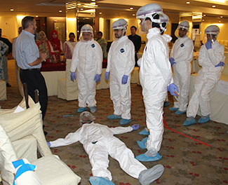 Workshop participants wearing hazmat suits and onlookers listen to a speaker in a conference room, actor lying on floor