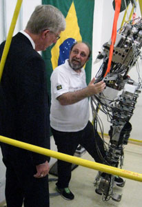 Dr Francis Collins observes as researchers demonstrates foot movement of a large metal robot, Brazilian flag hangs in background