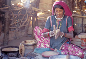 Woman sits cross-legged on ground rolling dough in hands, round flat bread cooking in pan on indoor cookstove