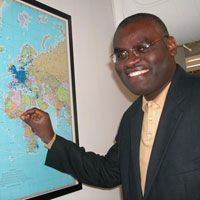 Dr. Clement Adebamowo looks at camera, smiling, points to Nigeria on a world map