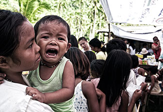 Crying child held while waiting in long line outdoors in the Philippines.