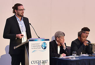 Male speaker in dress jacket speaks at a podium with sign that reads CUGH 2017, other panel members seated in background