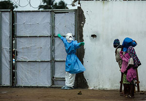 Healthcare worker outside Ebola clinic in full protective gear holds gate closed, warns away approaching people