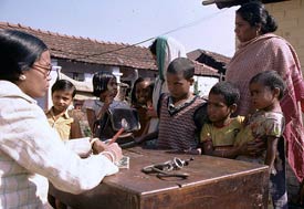 In Indian village, Woman sits writing, across table woman surrounded by six children waits and listens