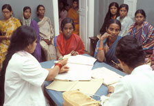 Two woman medical workers sit across table for two young Indian woman in saris, helping them fill out forms, many young women se