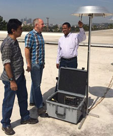 Three men stand next to equipment crate and tall silver metal tool measuring air quality
