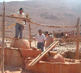 Three miners stand next to rough wooden supports and ramp, and dried red clay in front of dry, hazy hill