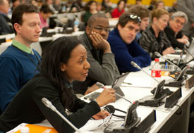 Woman attending large, busy conference session seated table speaks into a microphone to ask question from audience, many other a