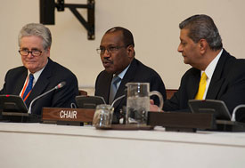 Three men seated at conference table in front of off camera audience, man in middle speaks