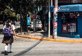 In Guatemala, a girl wearing backpack and school uniform crosses street toward market covered in ads for Pepsi and other product