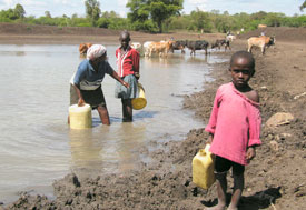 young girl in foreground carries plastic jug of water from muddy water source, woman and older girl in background fill jugs
