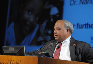 Ambassador Eric Goosby speaks at a podium at NIH, slide projected in background, picturing a young child