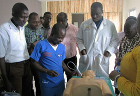 Medical worker in scubs demonstrates on dummy model laying on stretcher, on worker in white coat observes, large group observes