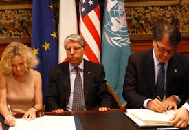 Dr Nora Volkow of NIDA, at table with two other men, signs documents, flags in background