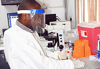 A scientist wearing a face shield and white lab coat works with samples in the lab