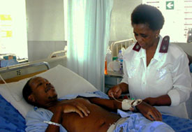 Man lies in hospital bed in gown in large room with other beds, medical worker attends him, observing his arm