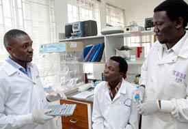 Three male medical workers in white coats wearing gloves, one explains to the other two