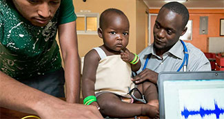 Medical worker examines young child who is sitting on an examining table in an indoor clinic setting, computer in foreground.