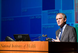 Dr. Christopher Murray speaks at a podium at NIH, slide of data visualization projected in background