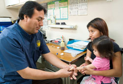 Male medical worker holds girl toddler's hand, mother holds the toddler in her lap, seated next to desk in clinic