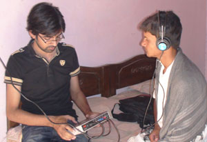 Young male researcher conducts hearing test on young man, who is seated on bed wearing earphone connected by chord to device