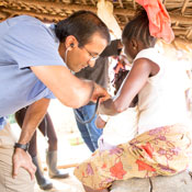 The photo shows Dr. Raj Panjabi, wearing a blue shirt and tan slacks, attending to a patient, a tween-aged girl in a white blouse and beige patterned skirt.  The photo was taken in the girl’s home and the background shows a thatched roof.