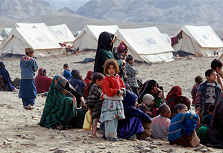 Group of Afghan refugees seated outdoors in a camp with tents in the background.