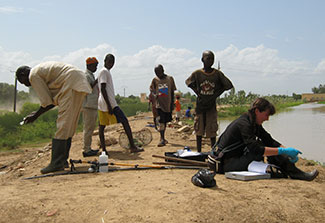 In Senegal, six researchers, some wearing gloves and boots, standing and seated on dry ground next to calm body of water.