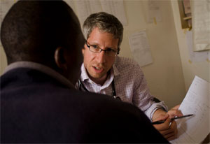 Dr Mark Siedner, wearing stethoscope, points to information on medical chart, speaks to patient seated opposite, back to camera