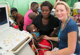 Dr. Amy Sims, cardiologist, examines a young child patient using cardiac equipment, woman holds child in her arms in busy clinic