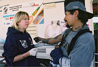 Woman wearing plastic gloves holds the arm (with many tatoos) of a man in front of a mobile health clinic.