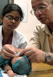 Older man places small plastic gadget on face of infant sized doll, younger female observes