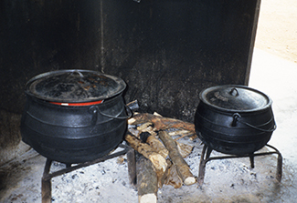 This photo shows two large black metal cooking pots on metal grates with a stack of wood between them.
