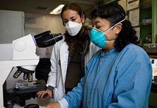 Two female researchers in surgical masks in lab work with microscope