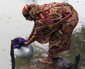 Woman dressed in sari stands on rock at edge of water, dips urn with sari cloth covering opening into water to filter it