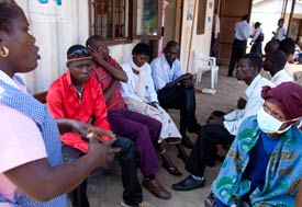 Woman standing wearing smock speaks to small group seated and listening to her, one seated woman wears medical mask